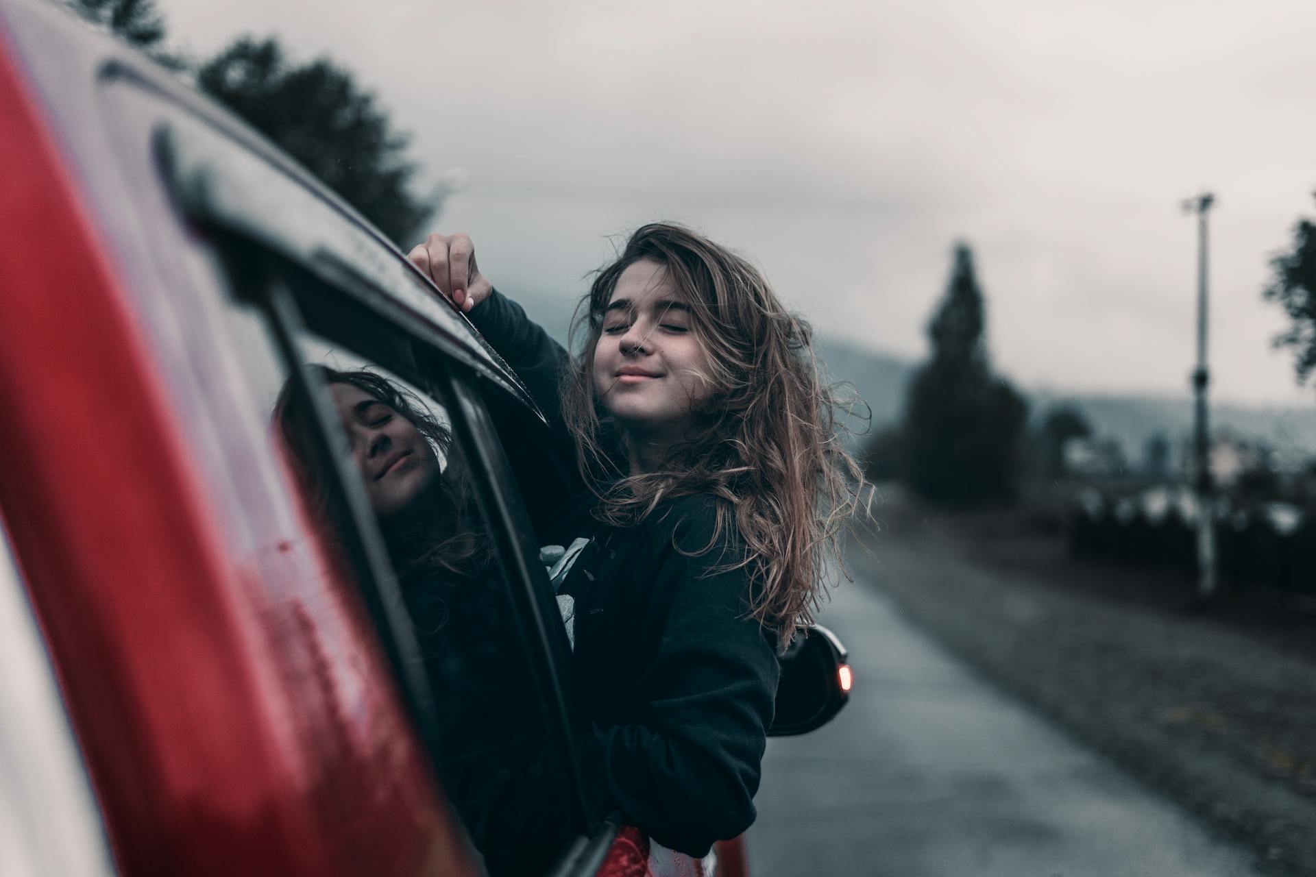 Side view of young dreamy ethnic female with closed eyes leaning out automobile window driving on asphalt road near trees and mountains under sky in countryside