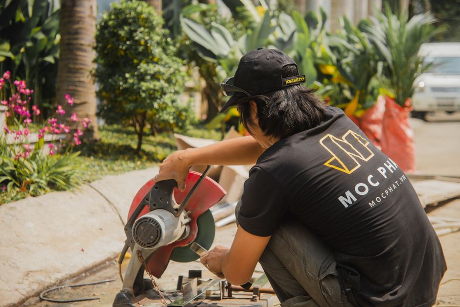 A carpenter uses a circular saw outdoors, surrounded by lush greenery in Hồ Chí Minh City.