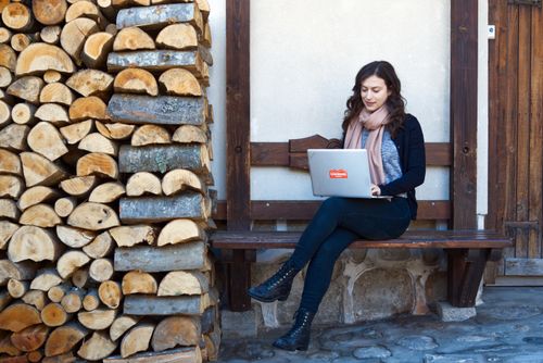 Free A woman sits on a wooden bench working on a laptop beside a stacked pile of firewood. Stock Photo