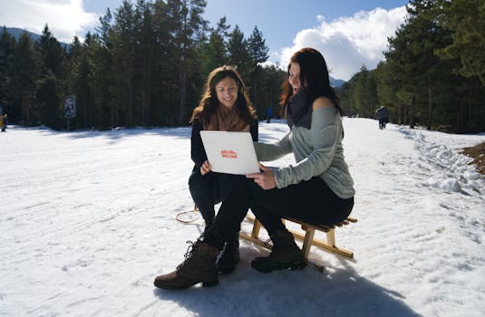 Two women working outdoors on a snowy mountain with a laptop.