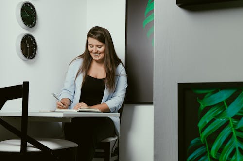 Cheerful young female journalist in casual clothes smiling while taking notes in planner during work in creative office
