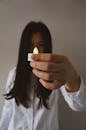 一位年轻的有色人种女性，留着深色长发，身穿白色衬衫，手拿小型无线人造蜡烛，站在一面模糊的墙壁前