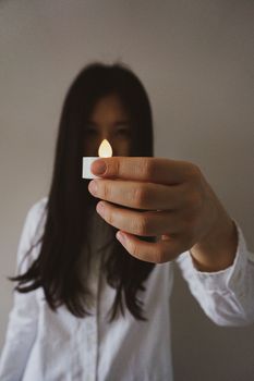 一位年轻的有色人种女性，留着深色长发，身穿白色衬衫，手拿小型无线人造蜡烛，站在一面模糊的墙壁前