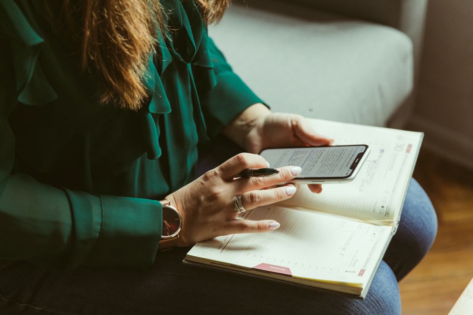 From above of crop anonymous female freelancer in elegant blouse using smartphone and writing information in notebook while working remotely at home sitting on sofa