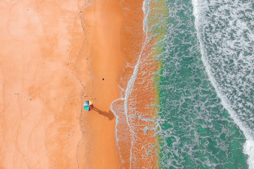 Amazing drone view of unrecognizable person with colorful umbrella resting on sandy beach near foamy ocean with turquoise water on sunny day
