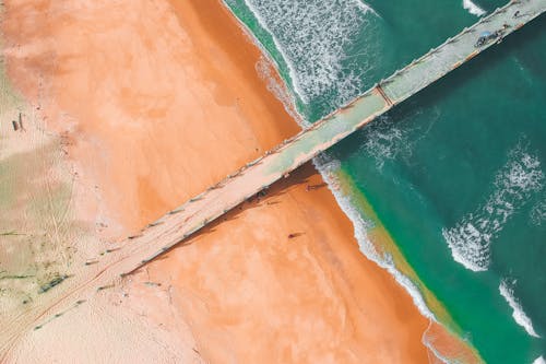 Breathtaking aerial view of long pier located on sandy coast of amazing ocean with turquoise water and foamy waves during sunny summer day