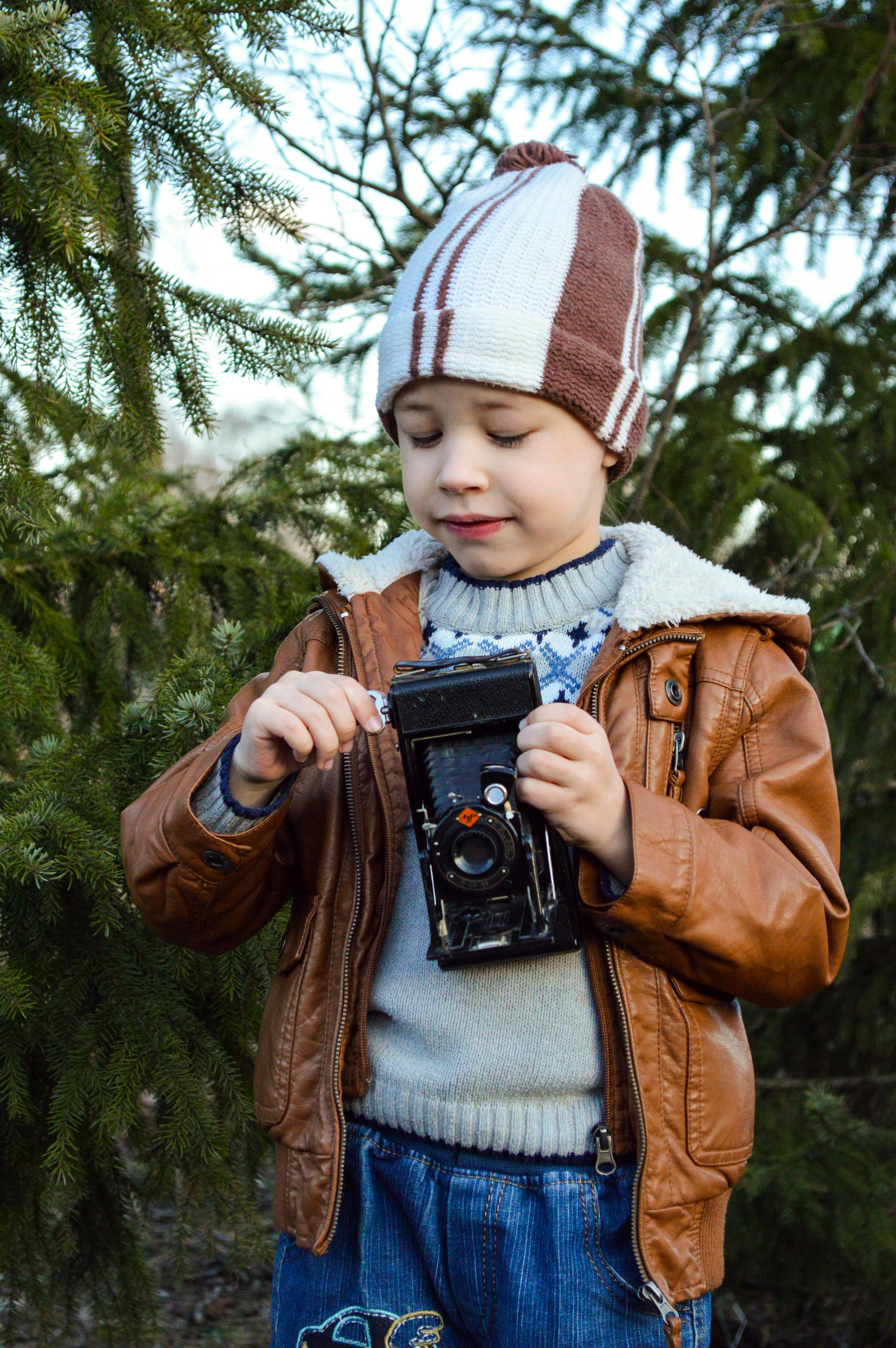 Joyful little boy in warm outerwear and hat playing with vintage film camera while resting in park with evergreen trees