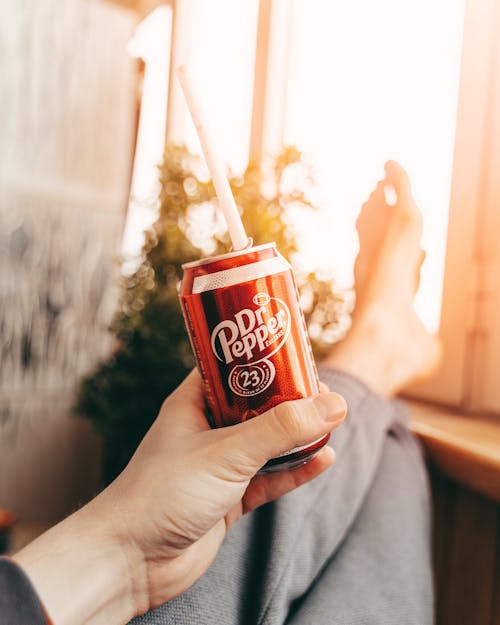 Crop anonymous person sitting with legs on windowsill and drinking refreshing soda from can with straw while relaxing at home