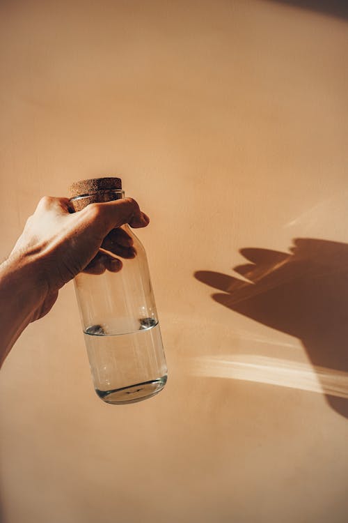 Free Crop anonymous person demonstrating transparent glass bottle of cold water near wall with shade on sunny day Stock Photo