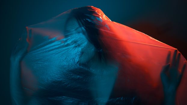 Unrecognizable female wrapped with translucent plastic bag in dark room with neon lights