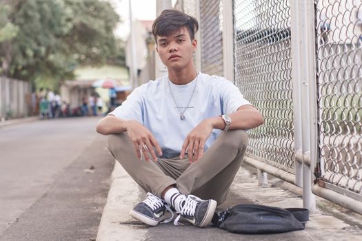 Full body of teenager in stylish outfit sitting on asphalt with legs crossed and looking at camera while resting next to chain link fence