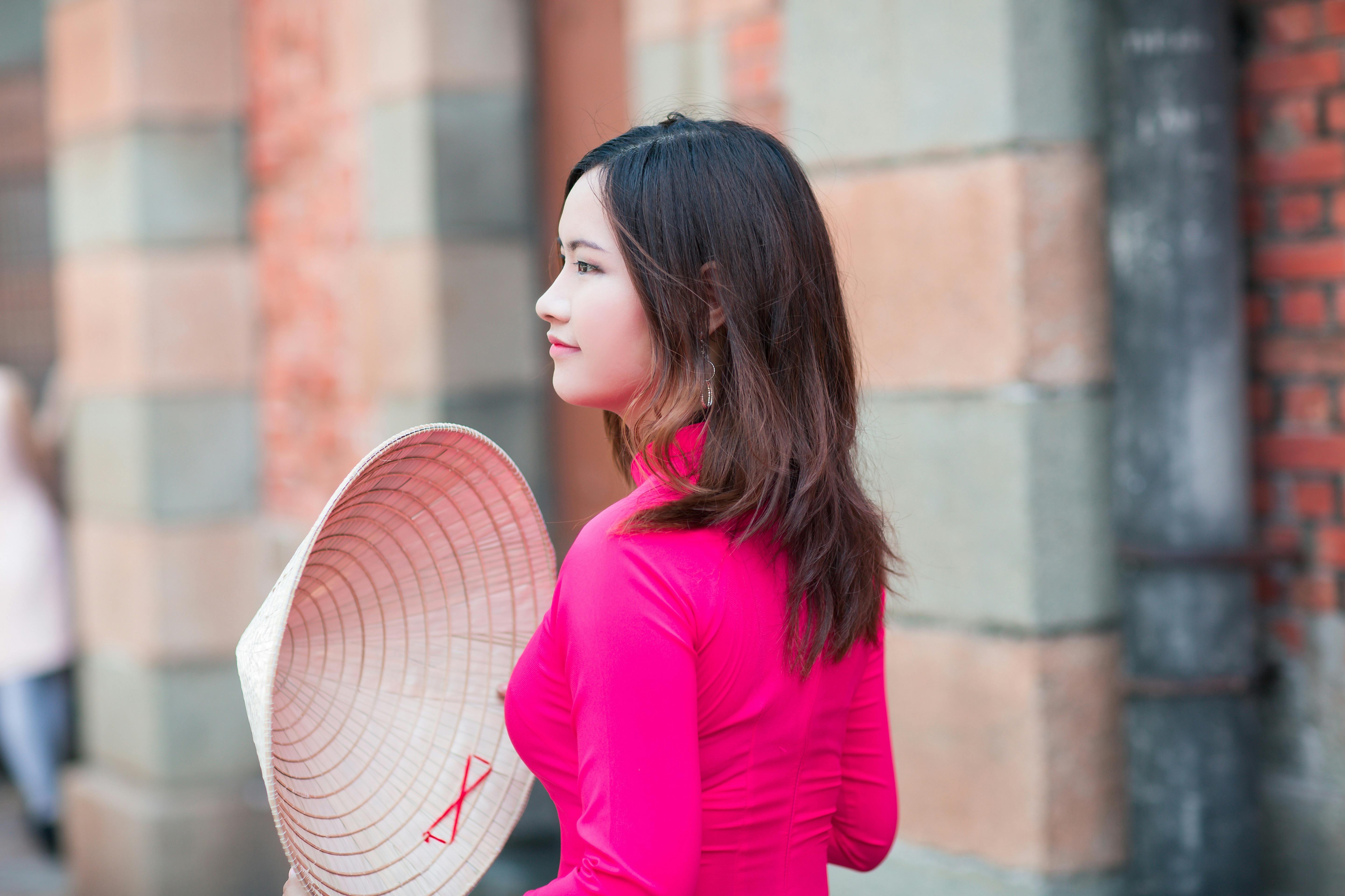 Free A graceful woman in vibrant pink traditional dress holding a conical hat outdoors against a brick wall. Stock Photo