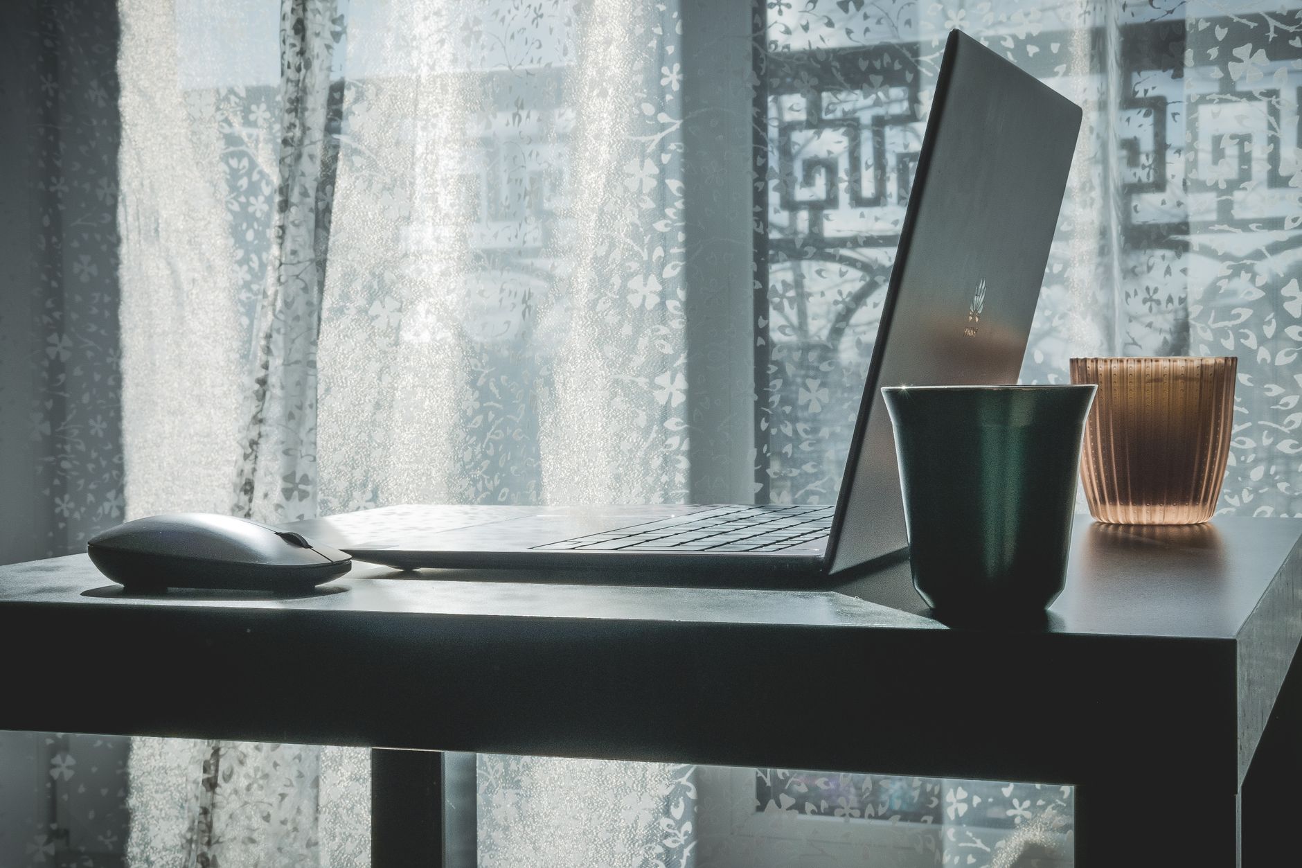 Cozy home office setup with a laptop, mouse, and coffee cup by a sunny window.