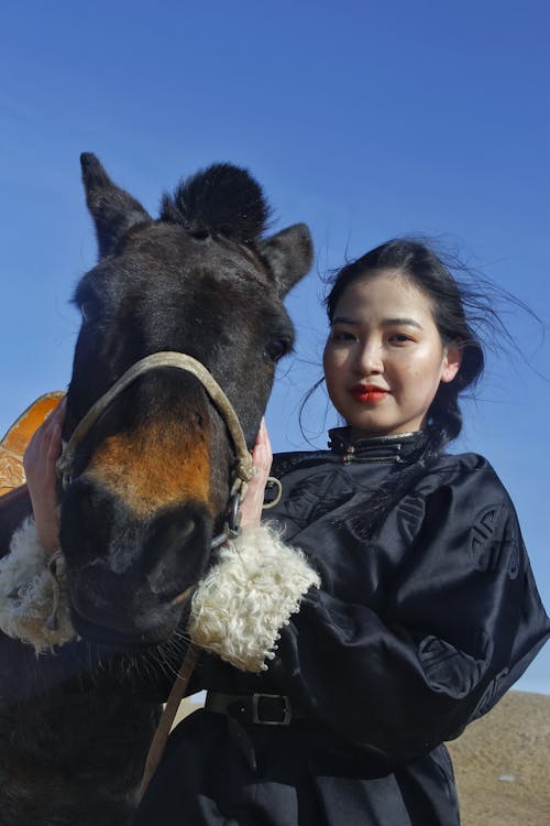 From below young content Asian female in black jacket caressing purebred adorable horse while standing on windy paddock