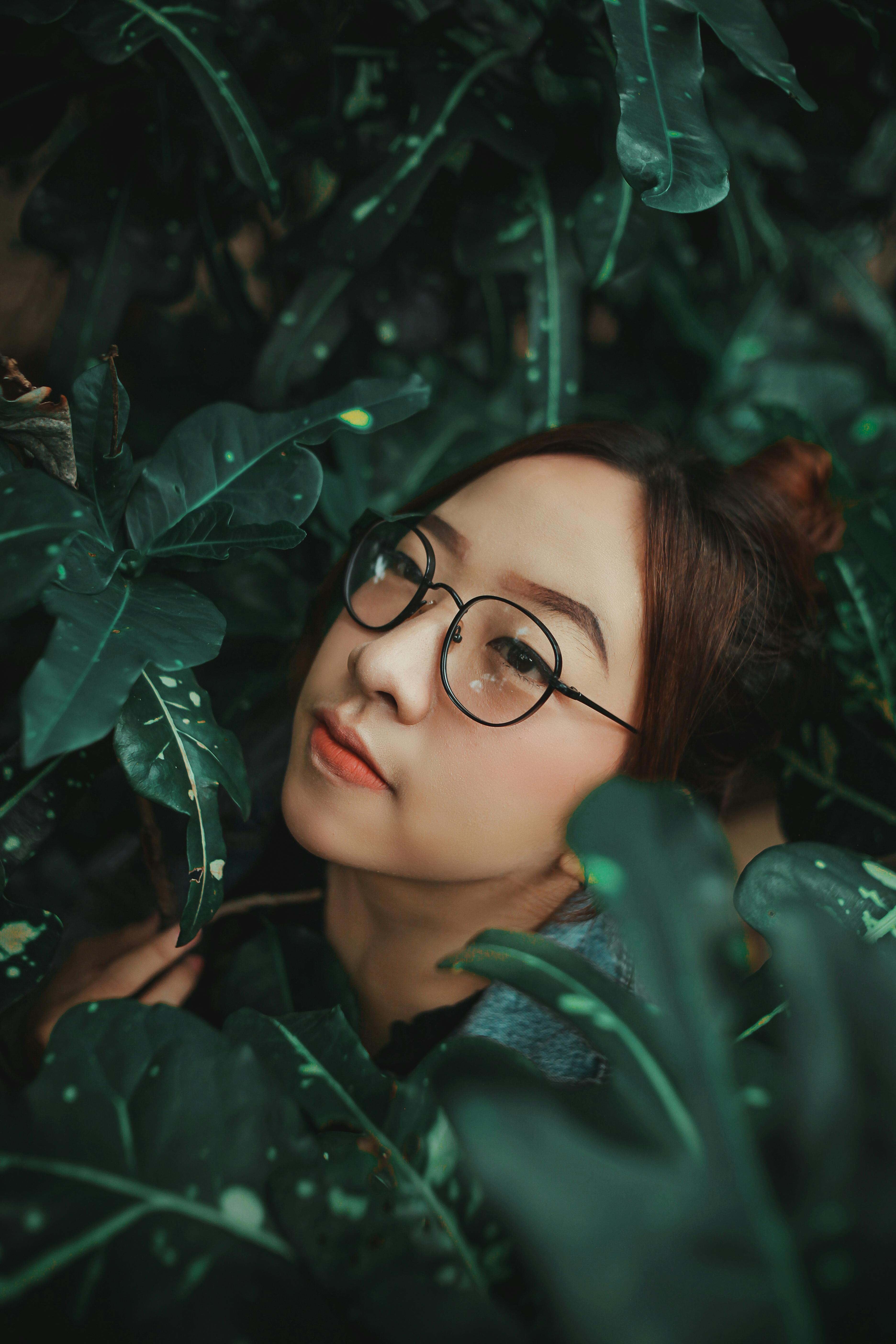 Portrait of an Asian woman with eyewear surrounded by lush green foliage, highlighting natural beauty and tranquility.