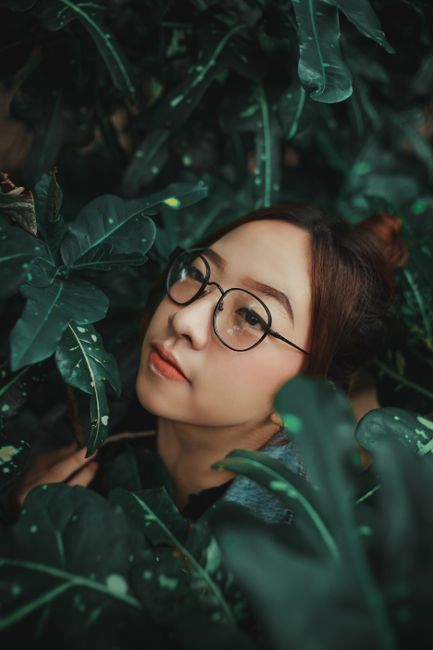 Portrait of an Asian woman with eyewear surrounded by lush green foliage, highlighting natural beauty and tranquility.