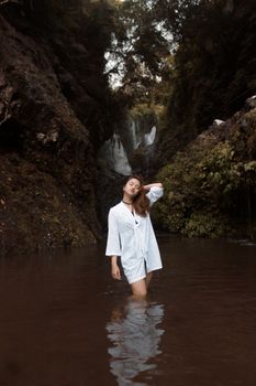 一位身穿白色连衣裙的女子在瀑布旁的宁静水池中放松身心，四周环绕着大自然
