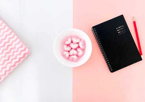 Notebook and pencil placed on white and pink desk with heart marshmallows