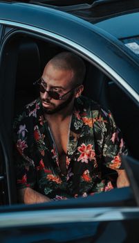 High angle of confident brutal bearded ethnic male in sunglasses and trendy shirt sitting in parked car with opened door and looking away
