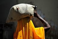 Back view of unrecognizable African female in traditional yellow headscarf carrying heavy sack on head while walking in village on sunny day