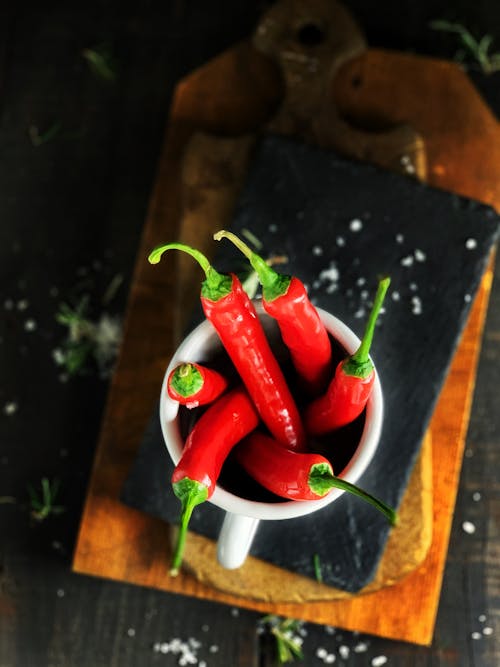 From above of heap of fresh red hot chili peppers in bowl placed on wooden cutting boards in kitchen