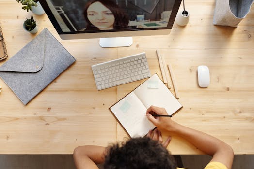 A teenager studying at home using a computer for online learning, taking notes in a notebook.