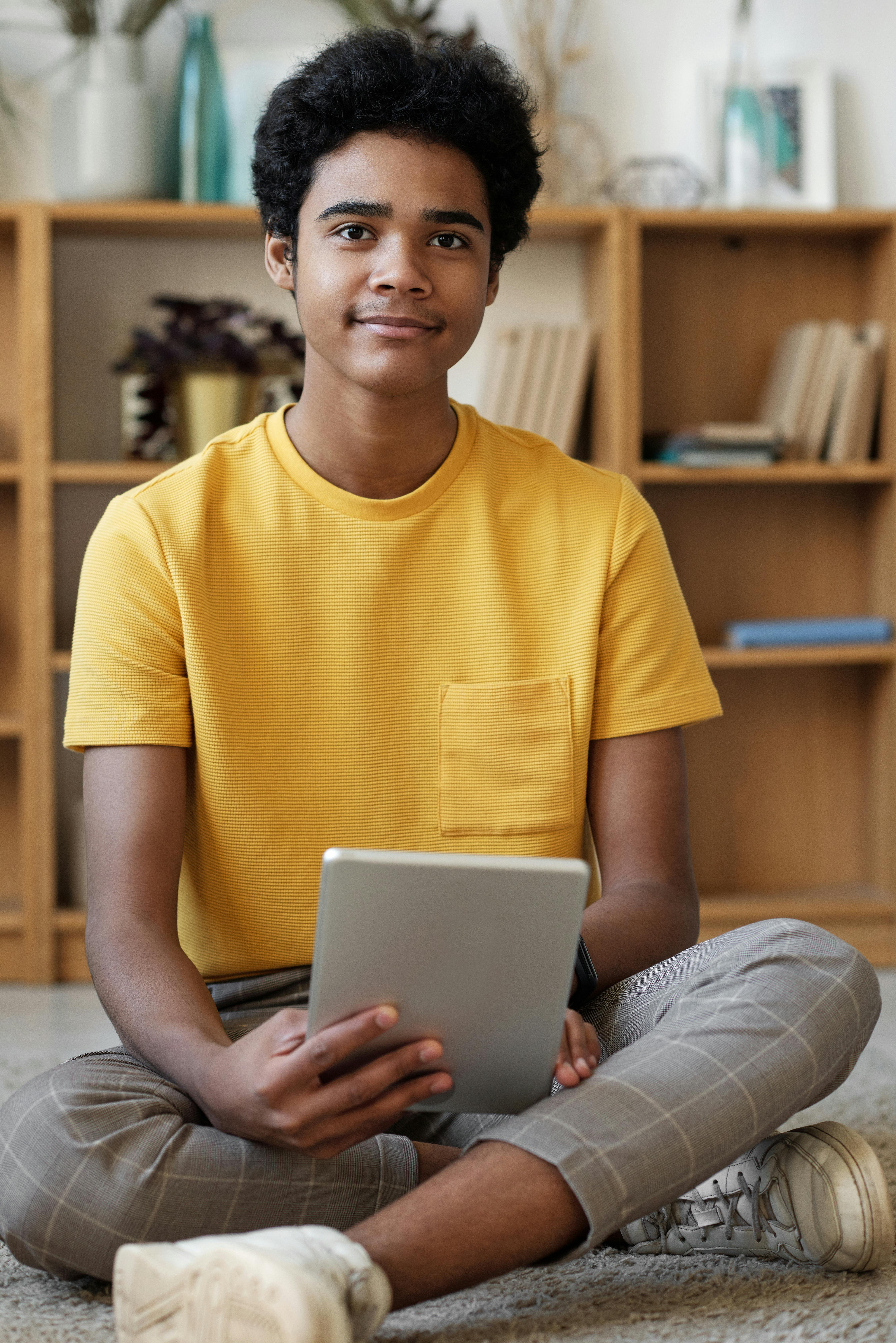 man in yellow crew neck t shirt and gray pants sitting on floor using silver tablet