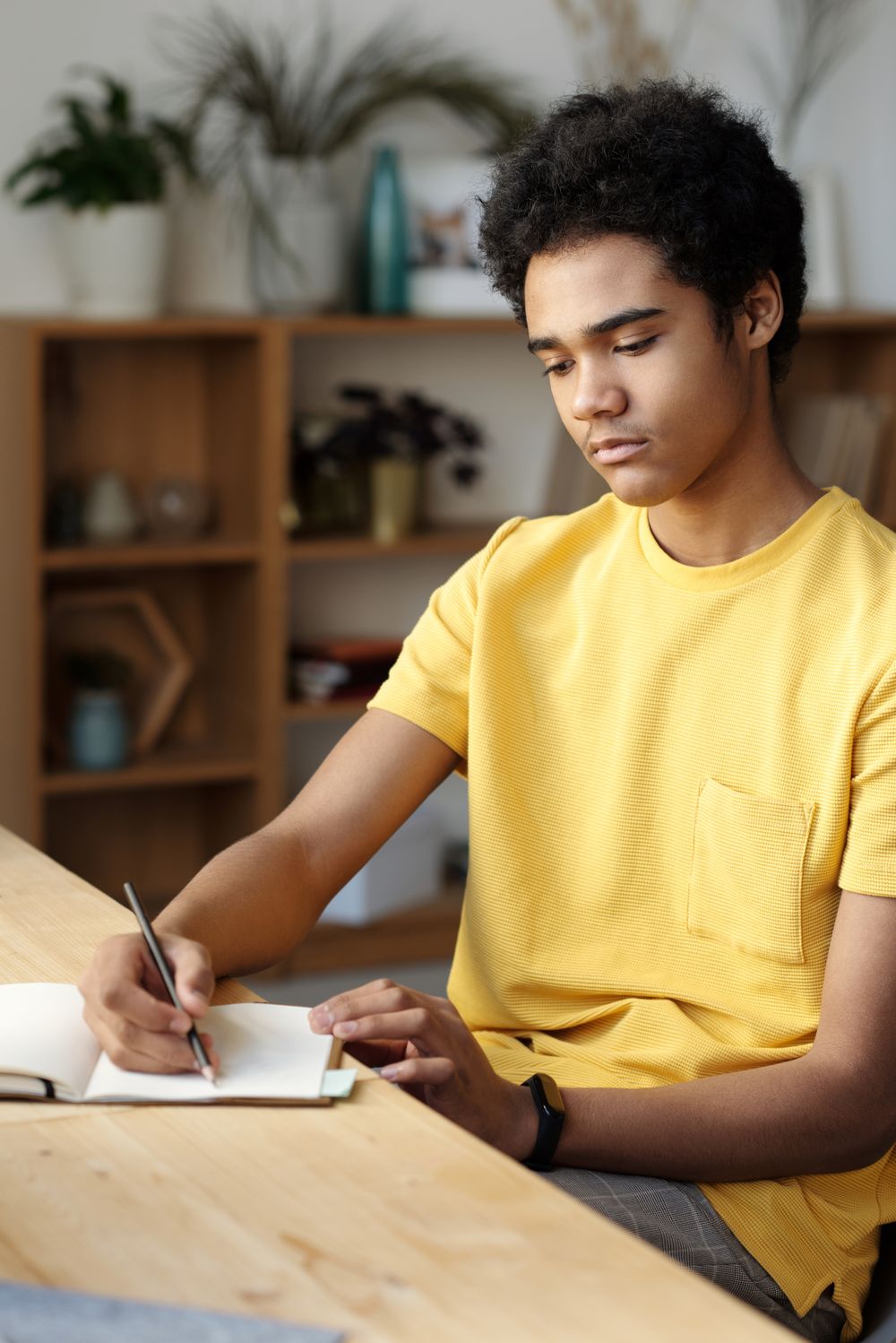 A high school student writing thoughtfully in a notebook at a wooden desk at home