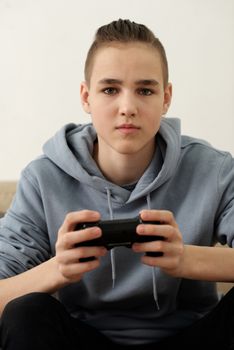 一个十几岁的男孩穿着灰色连帽衫，在室内全神贯注地玩着视频游戏机