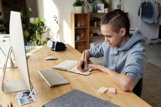 A teenage boy focuses on his studies at home, writing in a notebook during an online class.