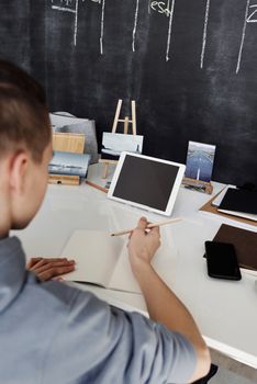 Teenager wearing gray hoodie studies with tablet and notebook at a white desk in front of a chalkboard.