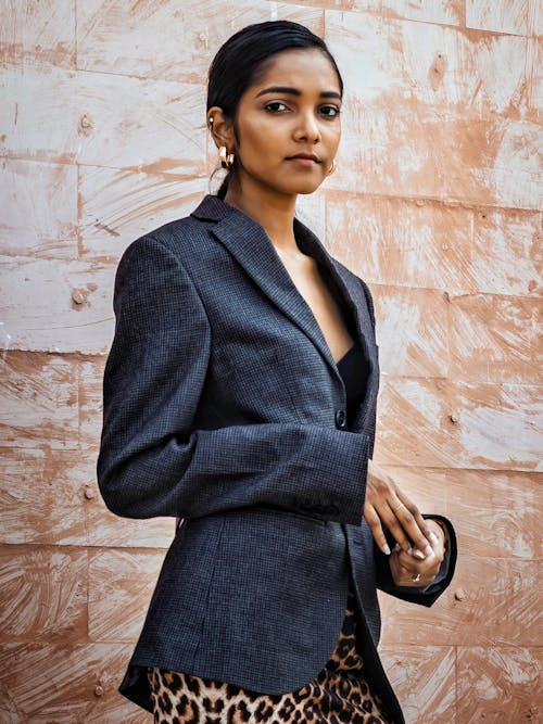 Serious ethnic female wearing classy jacket and dress looking away while standing against beige wall