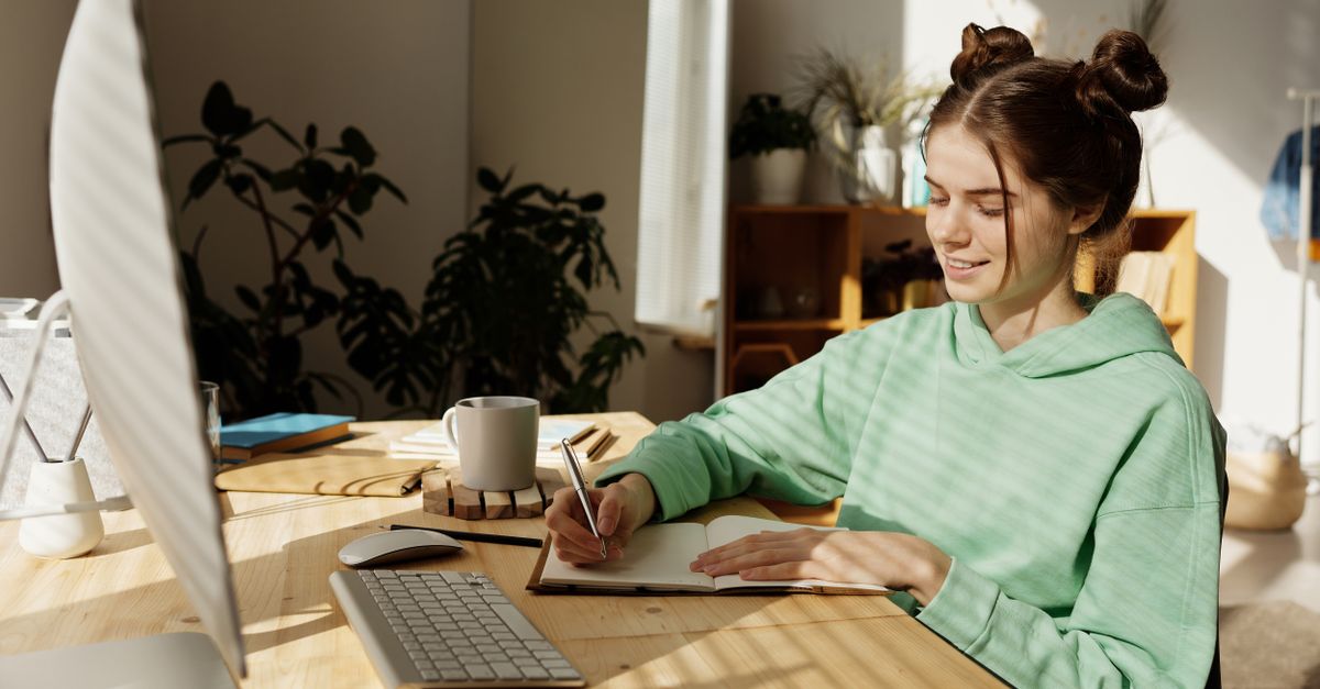 Teen girl in a green hoodie sitting at a desk, writing in a notebook, illuminated by sunlight.