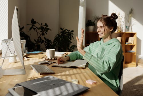 Free A Woman in Green Sweater Sitting Near the Wooden Table while Talking in Front of the Computer Stock Photo