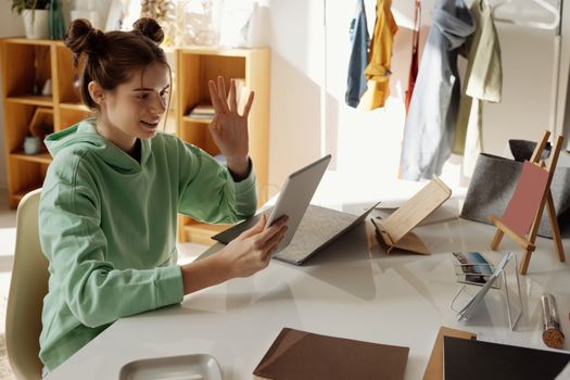 A young woman in a green hoodie video calls using a tablet in a cozy home office setting.