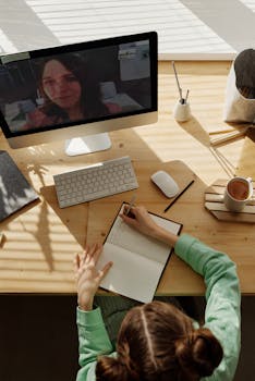 A teenager attends an online class from home, taking notes on a notebook during a video call.