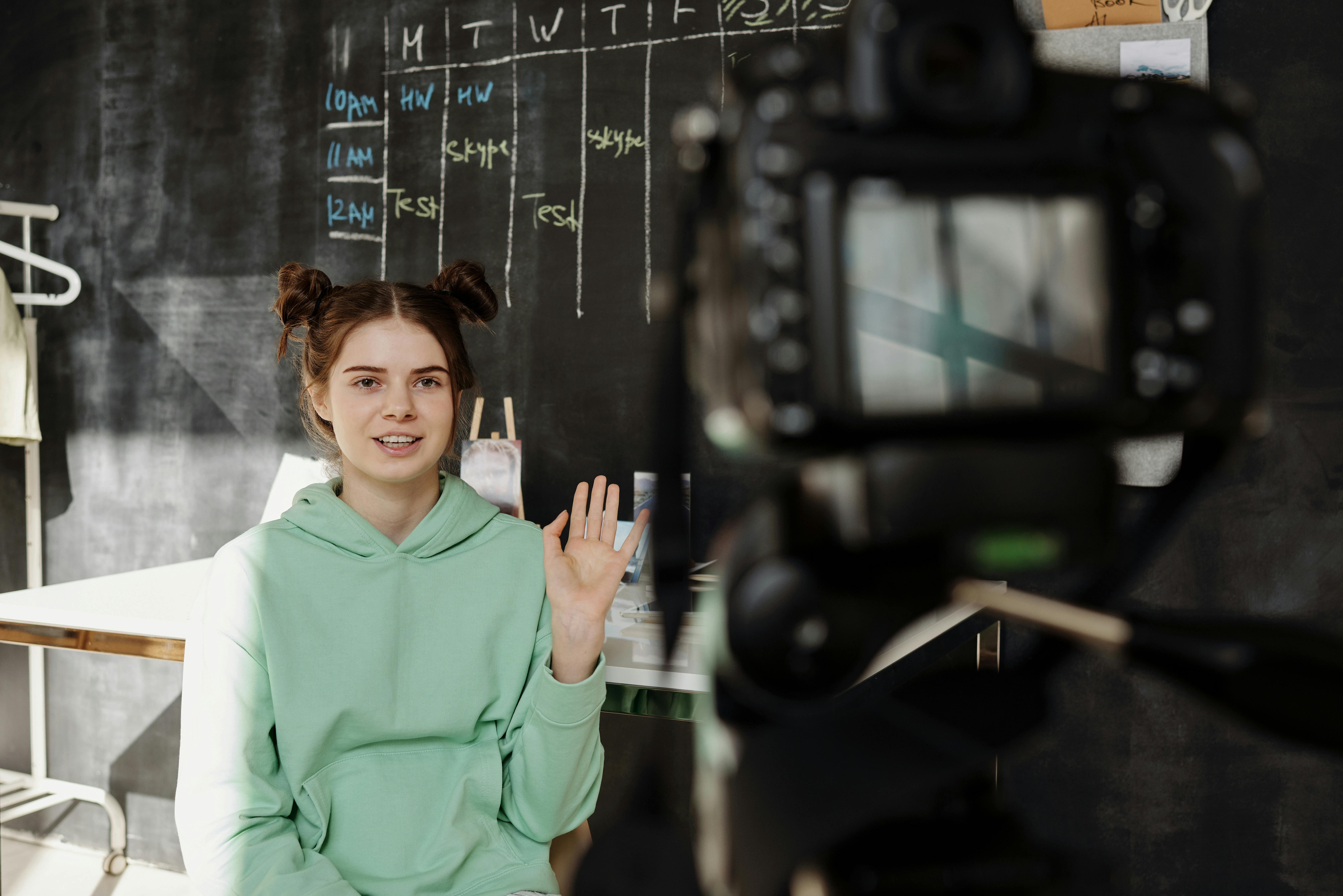 A person waving to the camera with a blackboard in the background