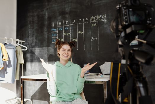 Young girl in a green hoodie smiling and gesturing during a video shoot in a creative home space.