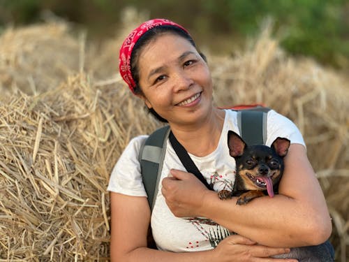 Cheerful ethnic woman with backpack smiling and embracing cute dog with tongue out