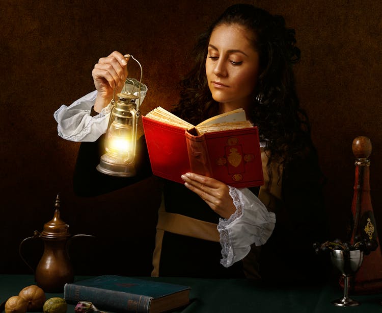 Focused Woman In Old Outfit Reading Book With Oil Lamp