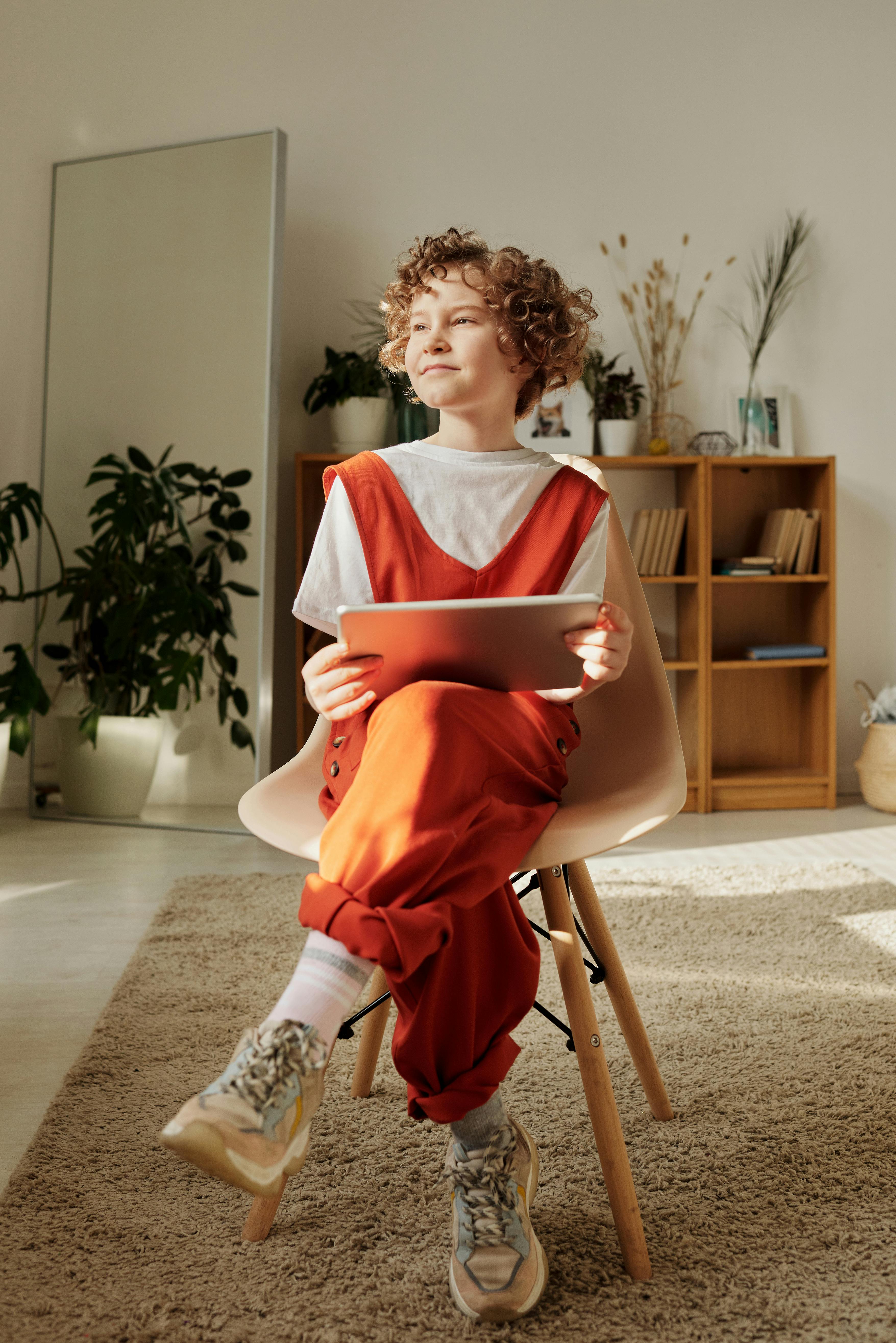 A young girl is sitting on a chair in her room while holding a tablet