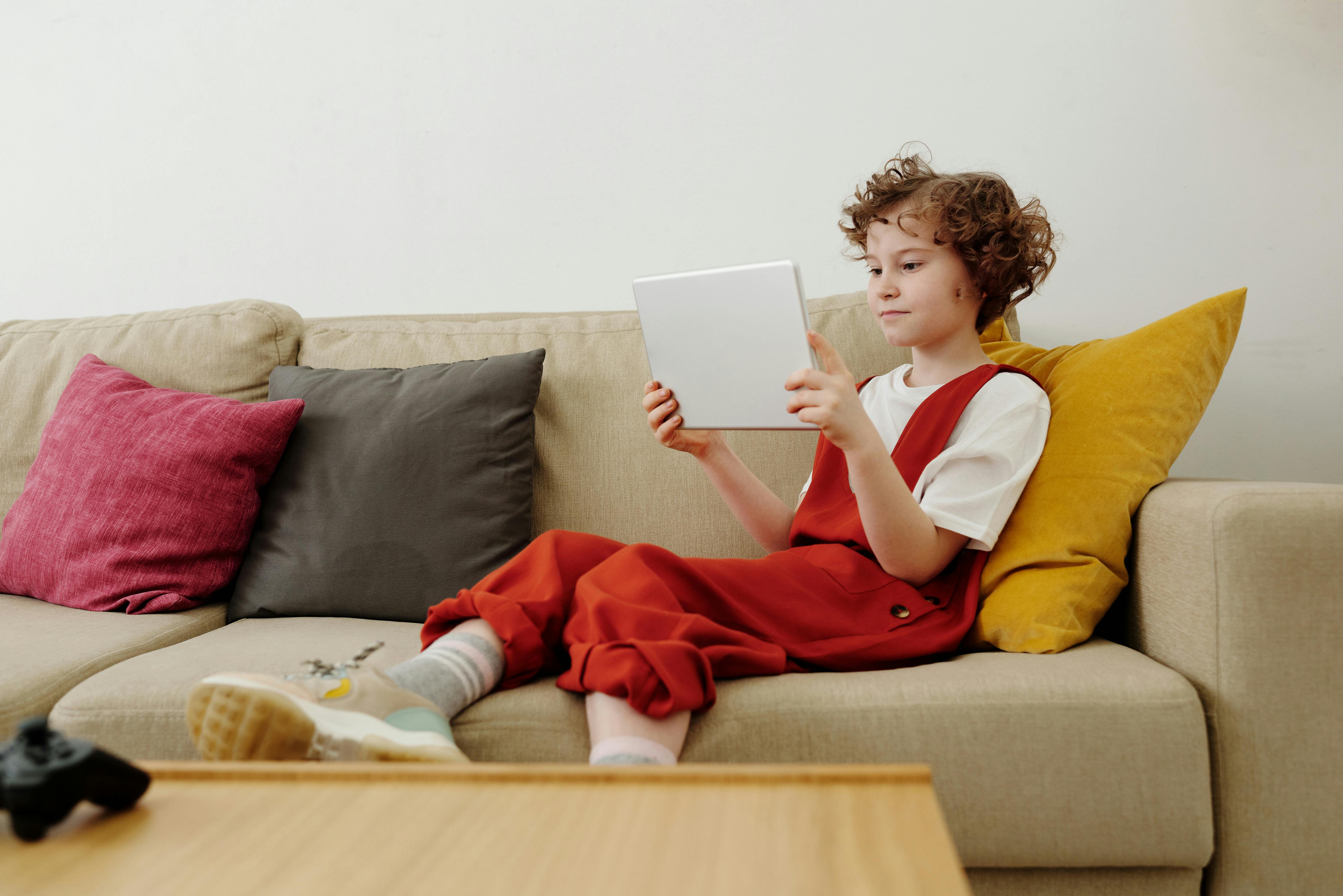 photo of child sitting on couch while holding tablet