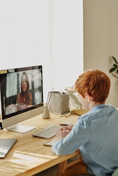 A young boy with red hair participates in an online class from home, using a desktop computer.