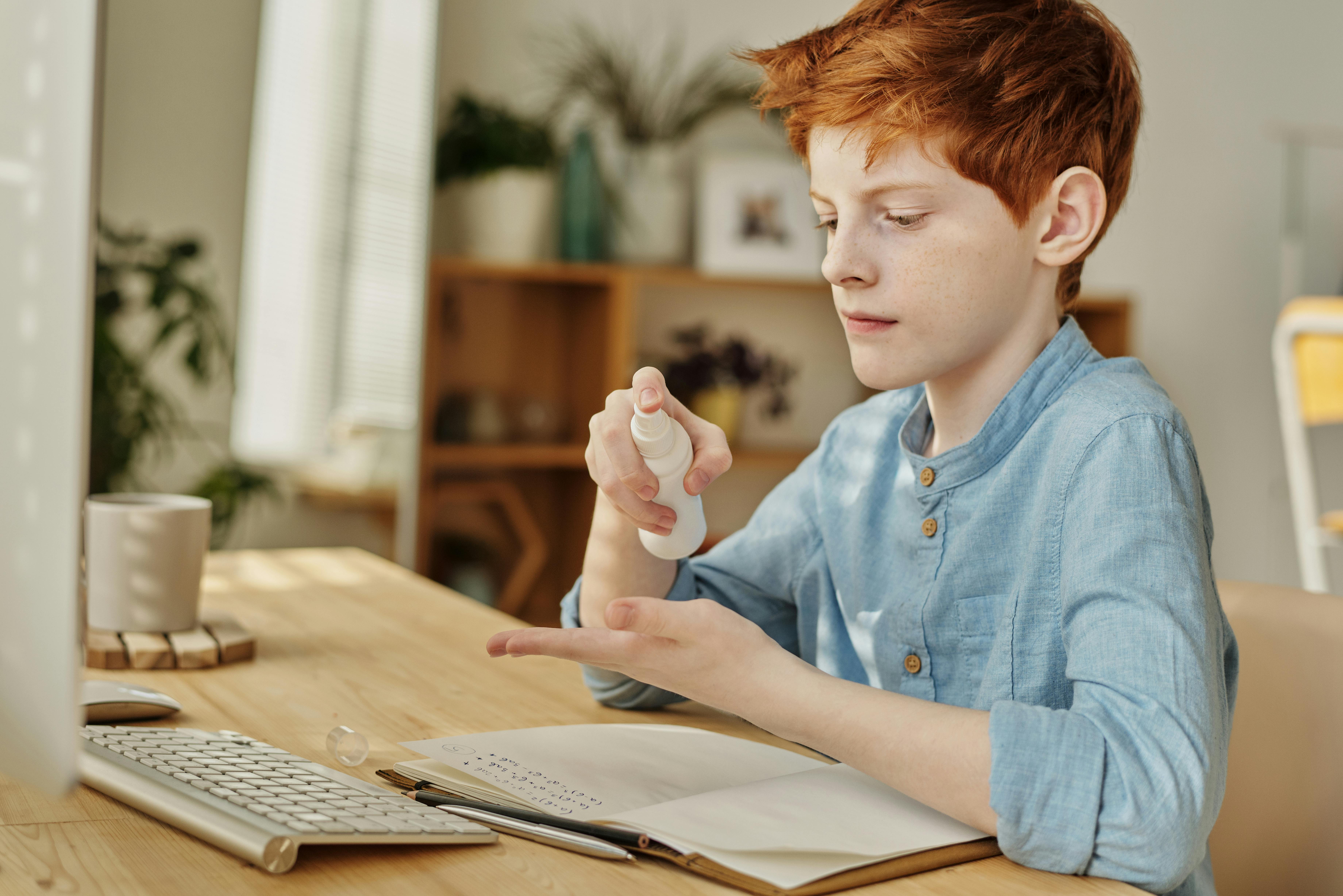 Red-haired boy applying hand sanitizer at a desk, emphasizing hygiene and safety.