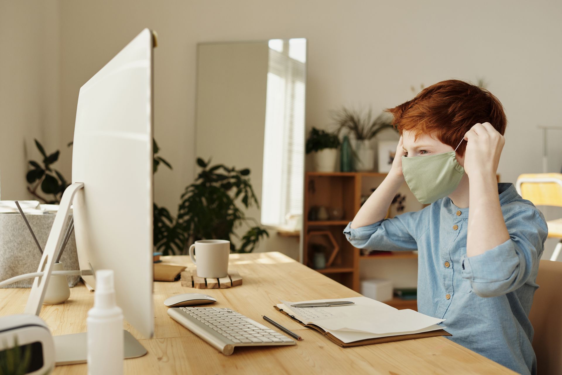 Child wearing a face mask while using a computer indoors during quarantine. Modern home office setup.