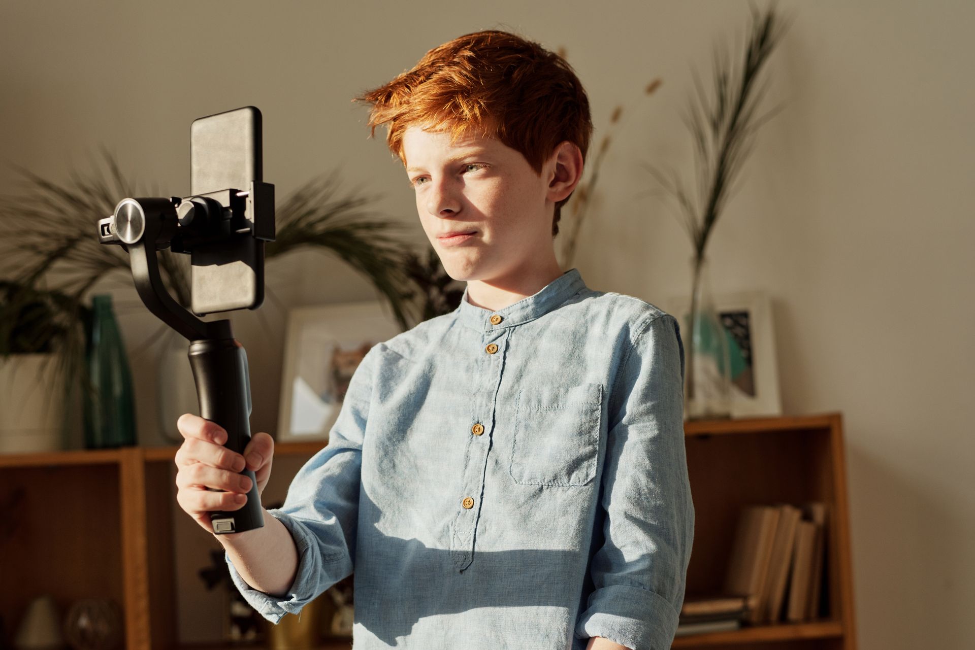 Young boy with red hair using a smartphone on a gimbal indoors, recording video.