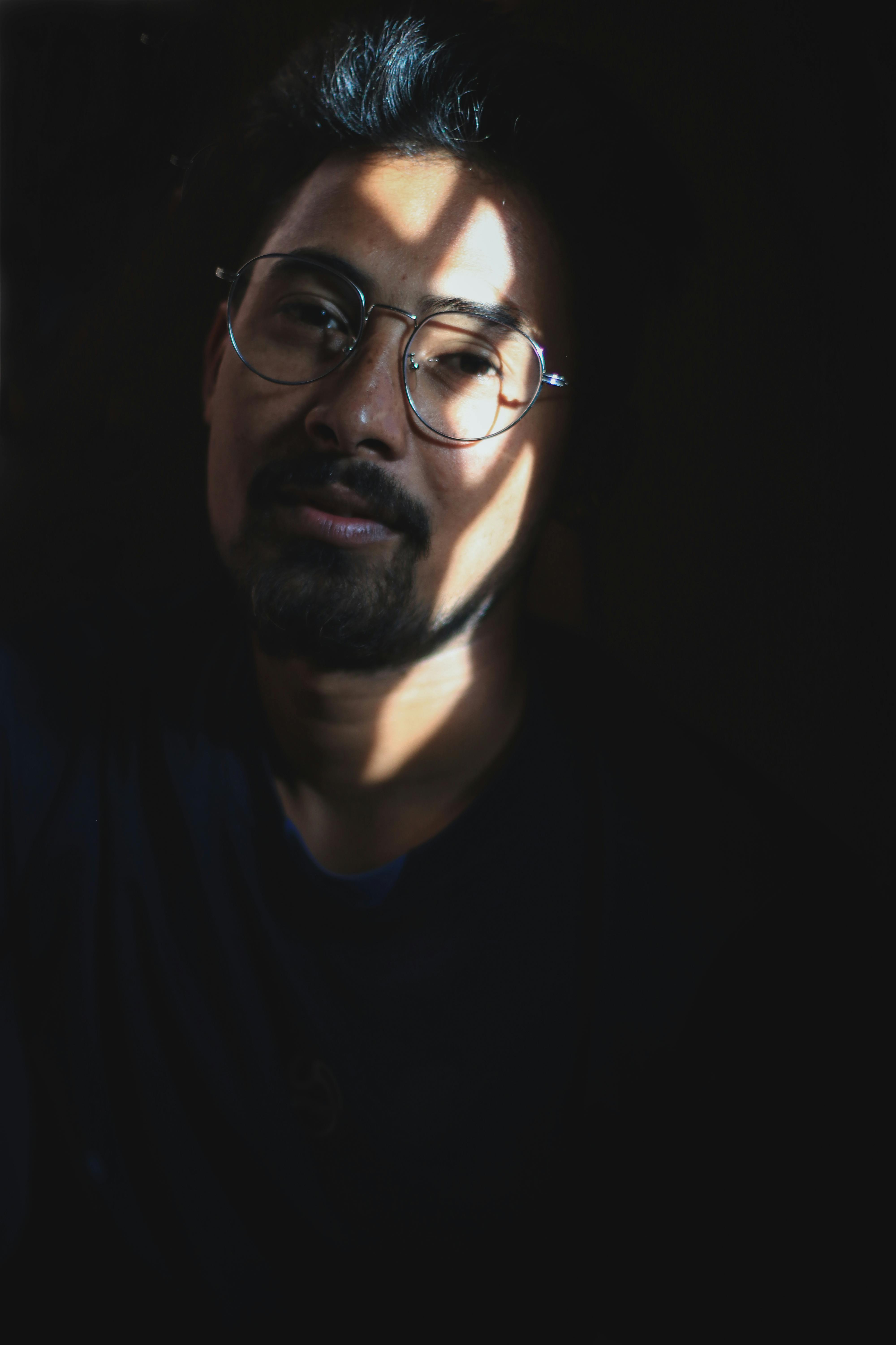 Close-up portrait of a bearded man wearing eyeglasses with dramatic shadows on his face.