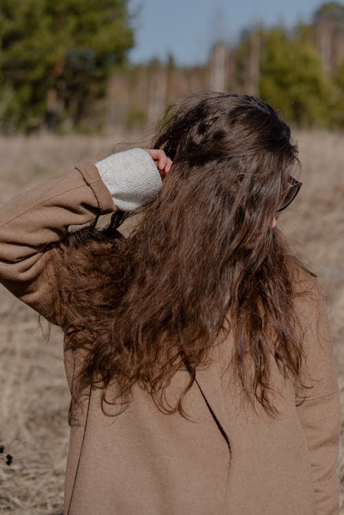 Unrecognizable female in warm outerwear and sunglasses standing in countryside and covering face with hair