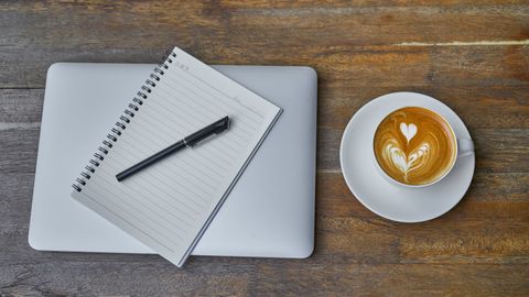 Flat lay of a coffee cup, notebook, pen, and laptop on a rustic wooden table.