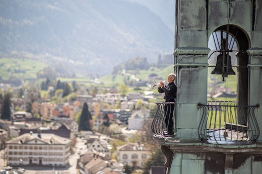 An elderly man plays a trumpet in a bell tower overlooking a scenic townscape.